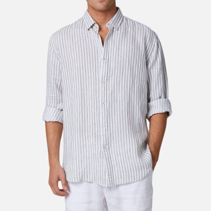 A man wearing the Industrie The Touville L/S Shirt—a light-colored, long-sleeve, button-up linen shirt featuring vertical gray and white stripes, with sleeves casually rolled up—paired with white pants. The background is plain and light.