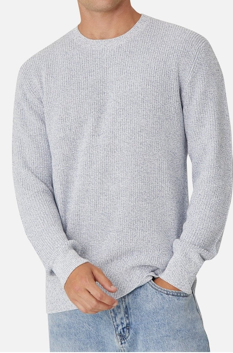 A man is wearing a light gray Industrie The Kingston Knit, paired with light blue jeans. He stands against a plain white background, casually placing one hand in his pocket.