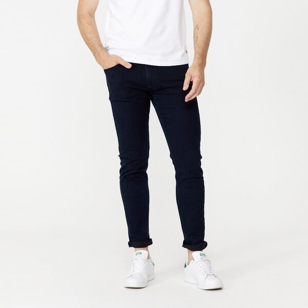A person is standing against a plain white background. They are wearing a white T-shirt, dark Levis 512 Slim Taper Jean, and white sneakers with green accents. Their hands are in their pockets and their face is outside the frame.