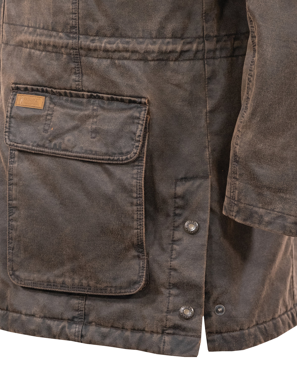 Close-up of a weathered brown Outback Nolan Jacket showing a large front pocket with a flap and a small label on the top left corner. The jacket features visible stitching and two metal snap buttons below the pocket. The rugged texture appears durable, making it an ideal water-resistant jacket.