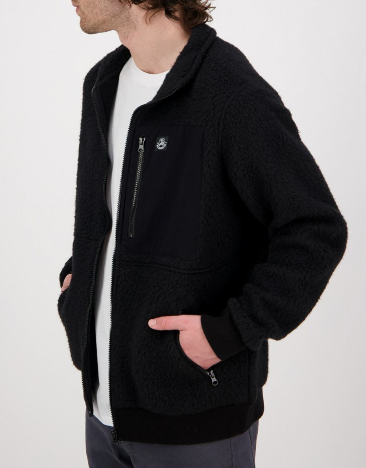 A person wearing a Swanndri Churchill Wool Fleece Jacket, featuring a black sherpa design with a zippered front, chest pocket, and small logo. Their hands are in the pockets as they turn slightly to the side against a plain white background.