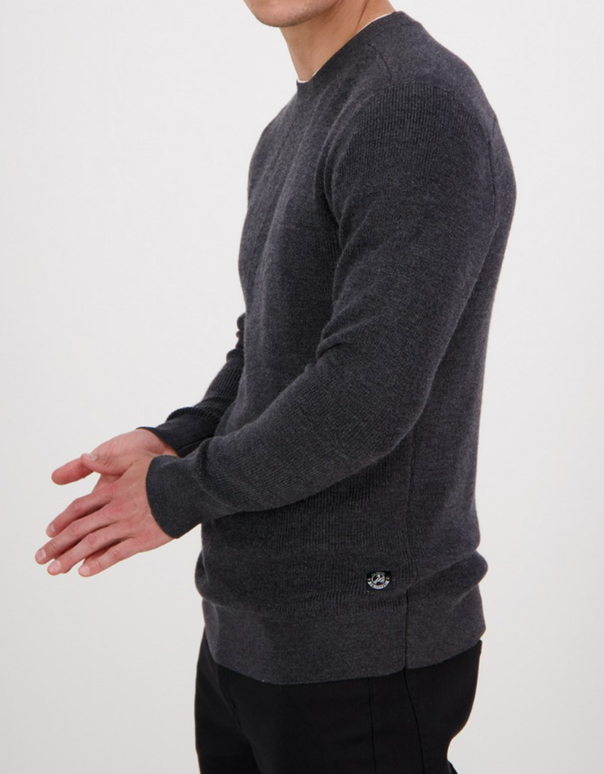 A person wearing the Swanndri Otahu River Crew Neck Merino Knit in dark gray and black pants stands against a plain white background, hands clasped and head slightly turned away—ideal for referencing our size guide or half chest measurement.