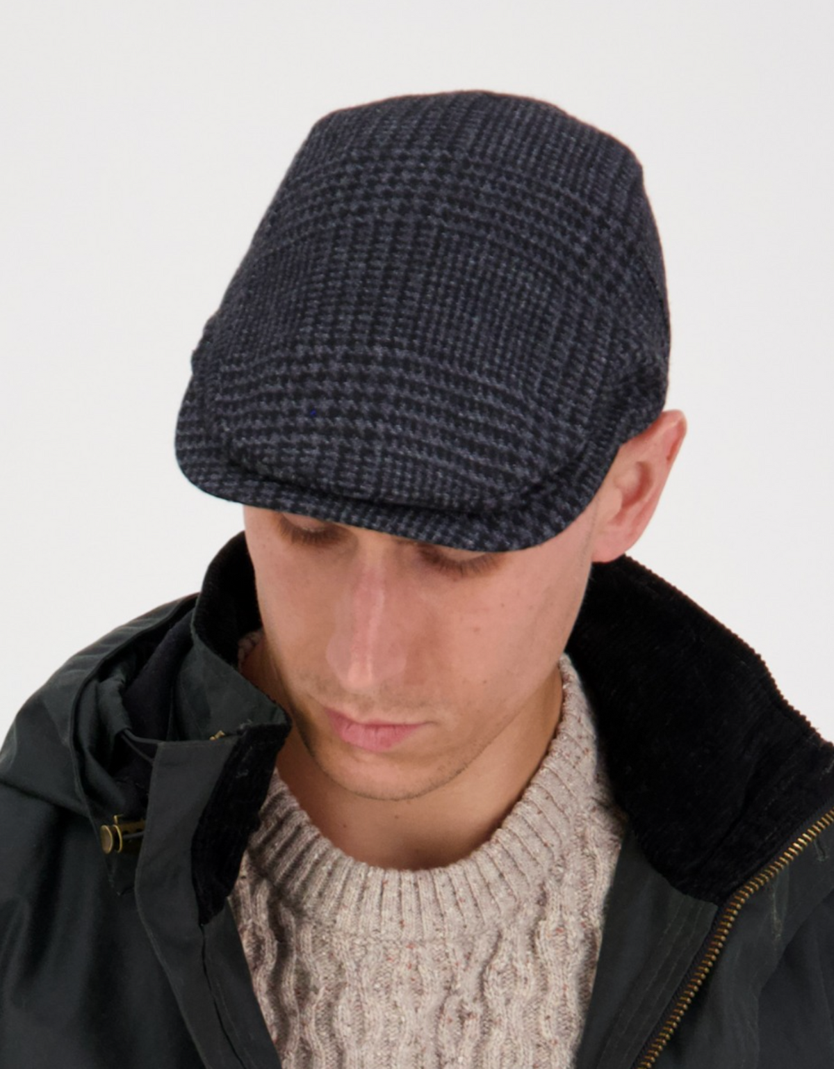Someone wearing the dark Swanndri Bristol Flat Cap and a dark jacket over a cream sweater exudes classic style, looking down against a plain white background.