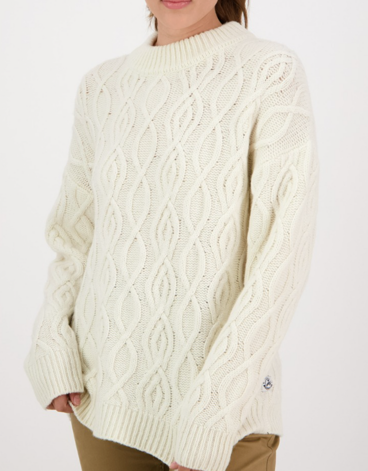 A woman models the Swanndri Pendeen Cable Neck Crew—a cream lambswool jumper with chunky knit, long sleeves, and a high neck—paired with beige pants against a white background.
