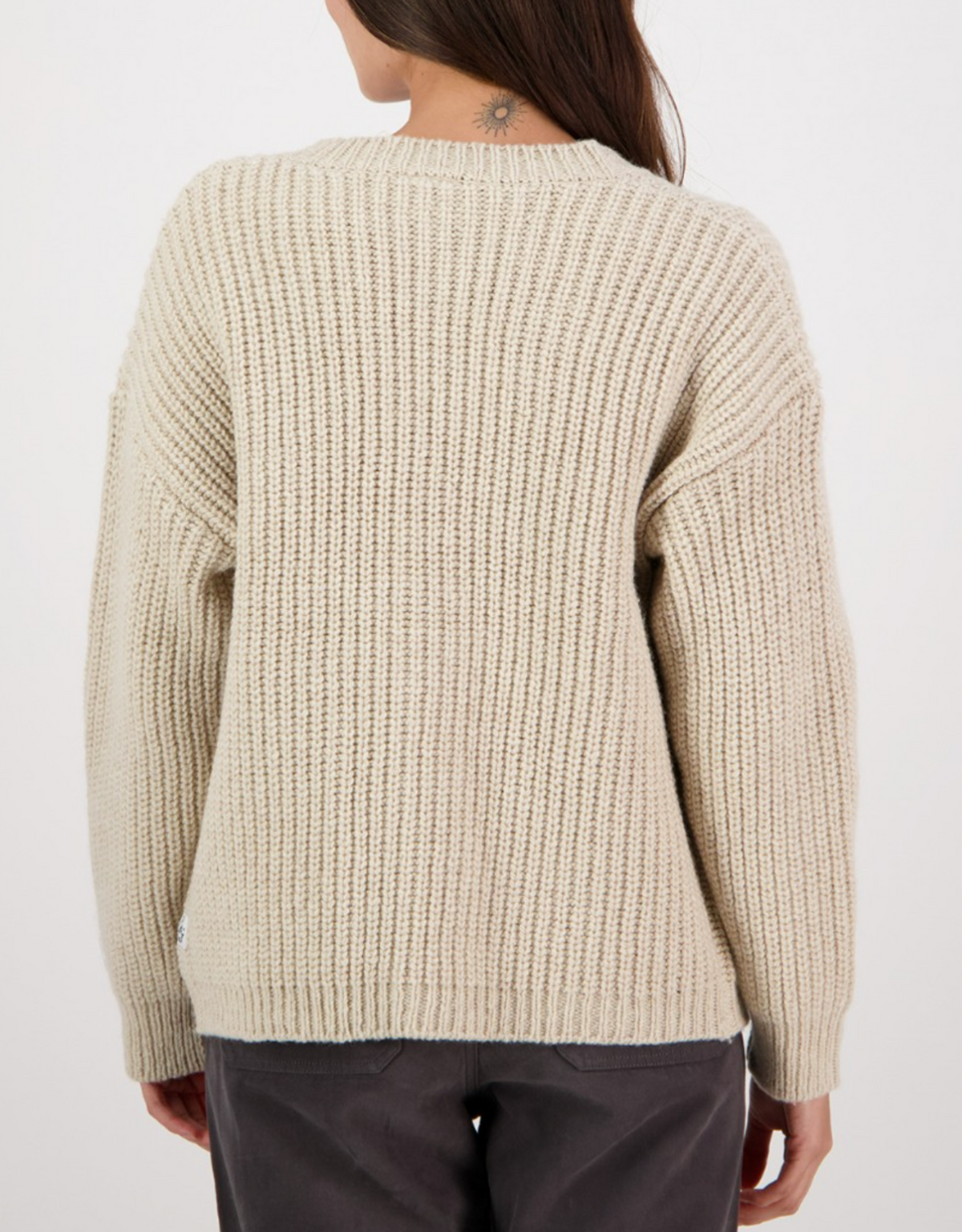 A woman with long brown hair, seen from behind, wears the Swanndri Braunton Knit Cardigan in cream and dark gray pants. A small tattoo is visible on the back of her neck.