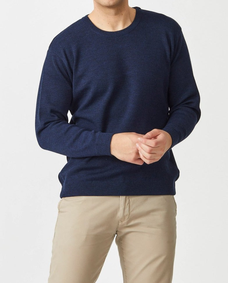 A person models the Aklanda Crew Pullover Australian Made in navy blue Merino wool with beige pants against a white backdrop, hands together in front, emphasizing the garment's exceptional quality without showing their face.