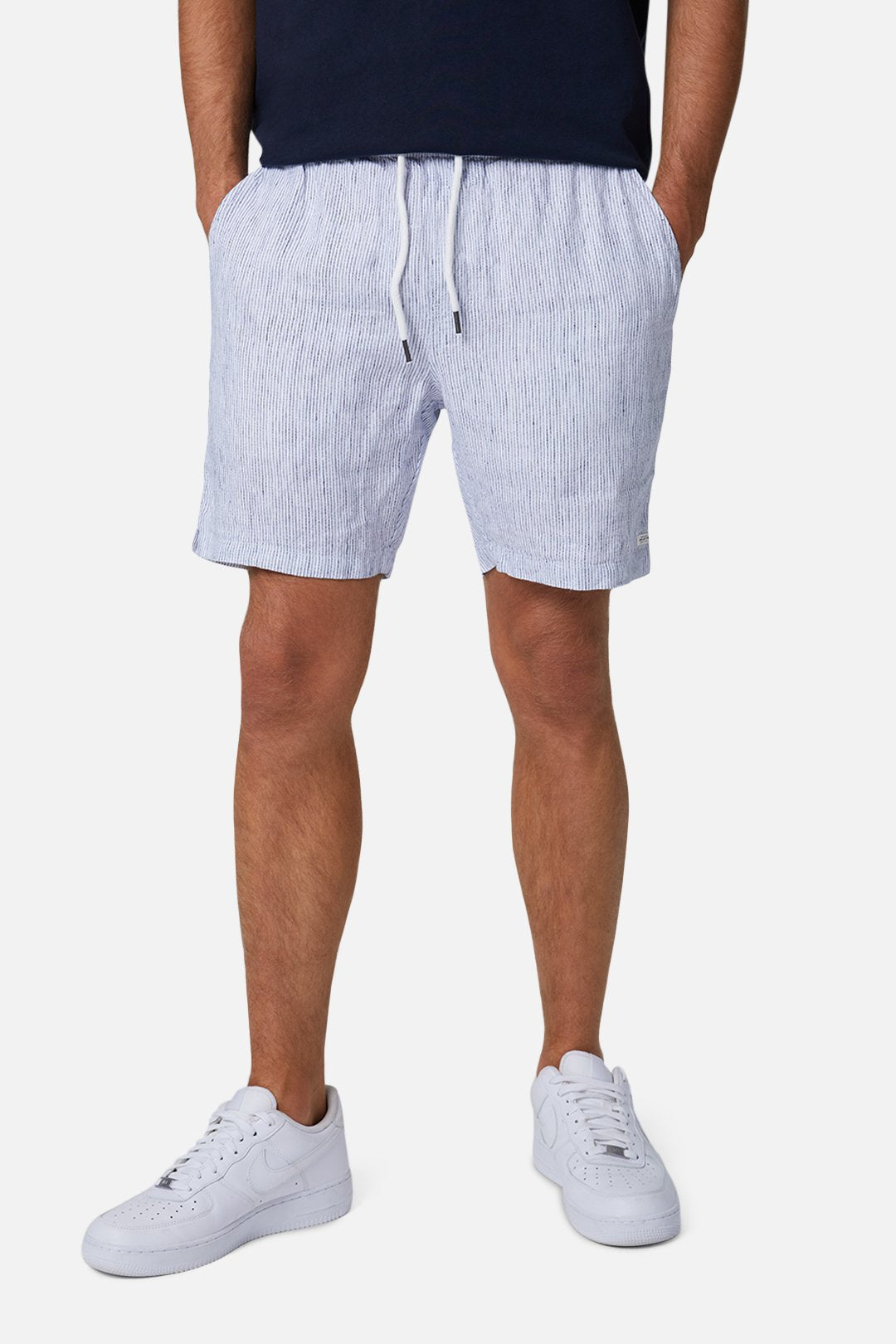 Wearing Industrie The Cristo Short in light blue and white stripes with an elastic waist, paired with a dark shirt and white sneakers. Hands rest in shorts' pockets, image cropped from the chest down against a white background.