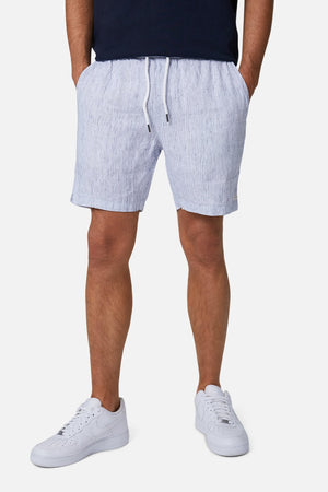 Wearing Industrie The Cristo Short in light blue and white stripes with an elastic waist, paired with a dark shirt and white sneakers. Hands rest in shorts' pockets, image cropped from the chest down against a white background.