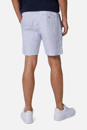 A person wears Industrie The Cristo Short in light blue and white stripes, made of 100% linen with an elastic waist, paired with a dark shirt and white sneakers. They are seen from the back against a plain white background.