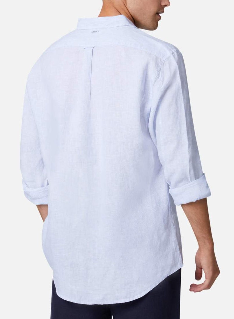 A person stands with their back to the camera, wearing the Industrie The Rochecroft Shirt, a light blue long-sleeved button-up made from 100% linen. The sleeves are rolled up to the elbows, giving it a soft, casual look. The comfortable shirt is paired with dark pants against a plain white background.