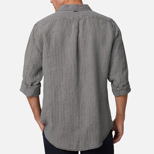 A man stands facing away, wearing the Industrie The Cilento L/S Shirt—a relaxed fit gray and white vertically striped linen shirt with rolled-up sleeves—paired with dark pants, against a plain light background.