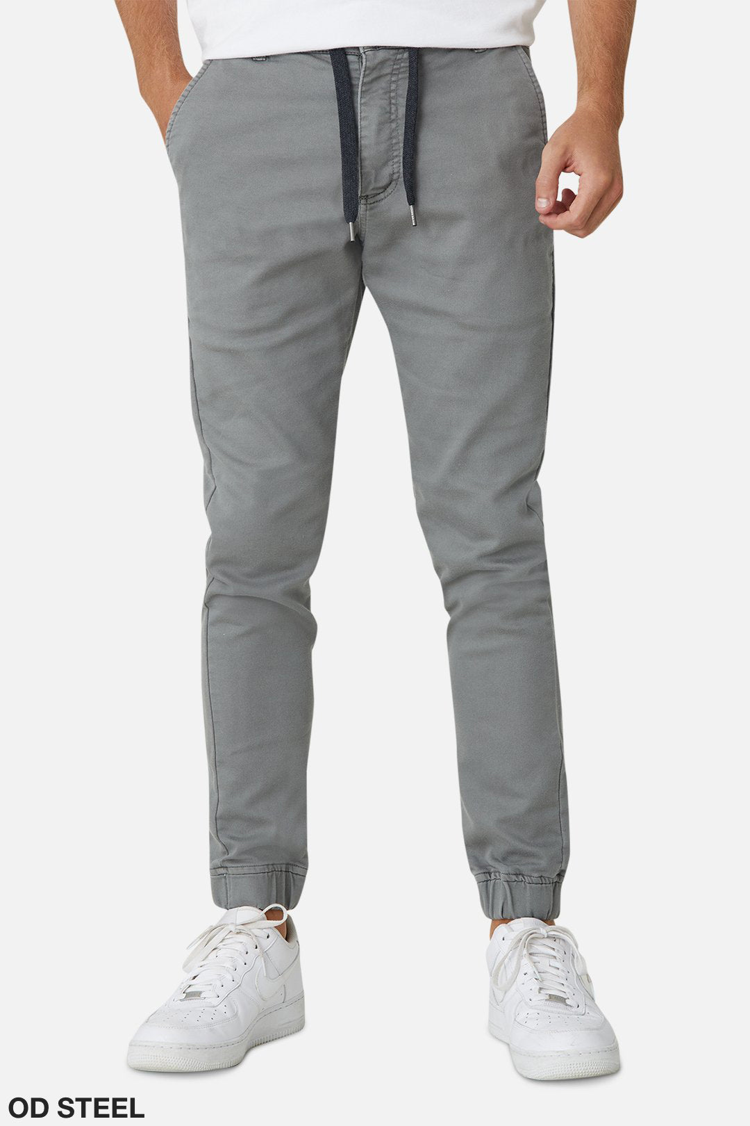A person's lower body in grey Industrie The Drifter Chino Pants with an elastic waist, hands in pockets, and wearing white sneakers. The drawstring and tapered design stand out against the white background.