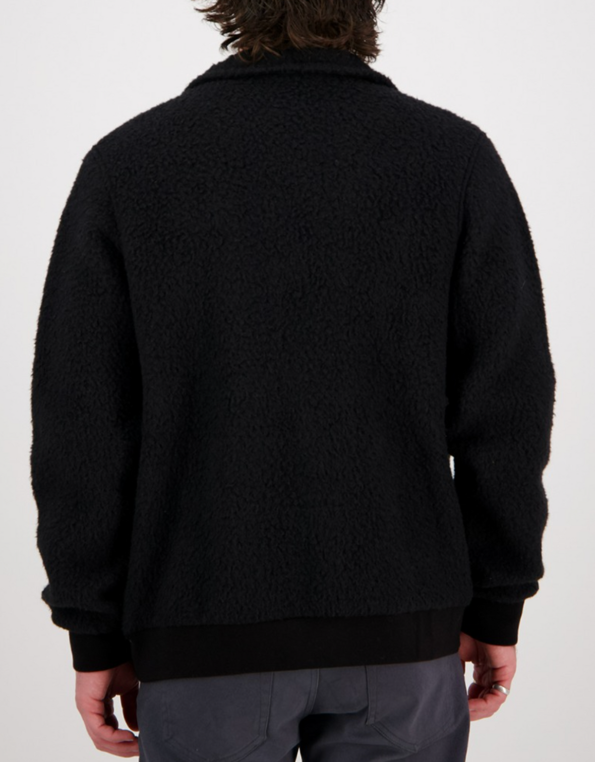 Someone wearing a Swanndri Churchill Wool Fleece Jacket in black stands facing away, with curly hair enhancing their casual look of dark-colored pants. The backdrop is a plain white.