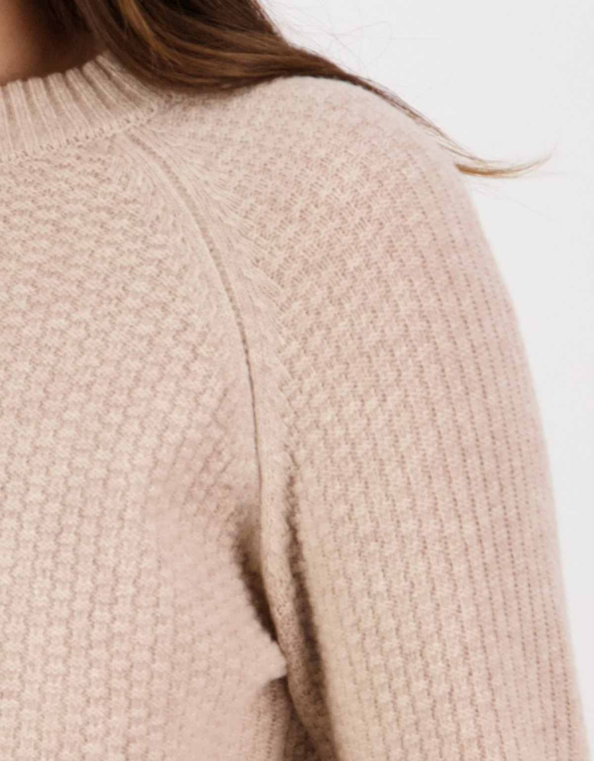 Close-up of someone wearing a Swanndri Hayes Park Waffle Knit Crew, a textured beige lambswool jumper with a ribbed collar. The soft fabric displays a knit pattern, and brown hair is partially visible over the shoulder.