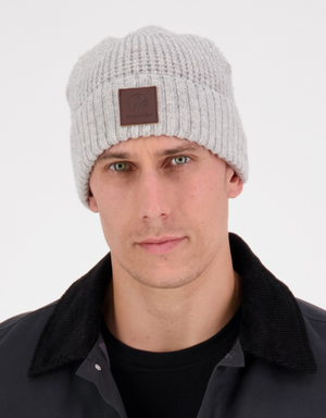 The person wears a light gray Swanndri Newgale Beanie with a waffle knit design and a brown patch on the front. They have a serious expression, pairing the beanie with a dark jacket featuring a black collar against a plain white background.
