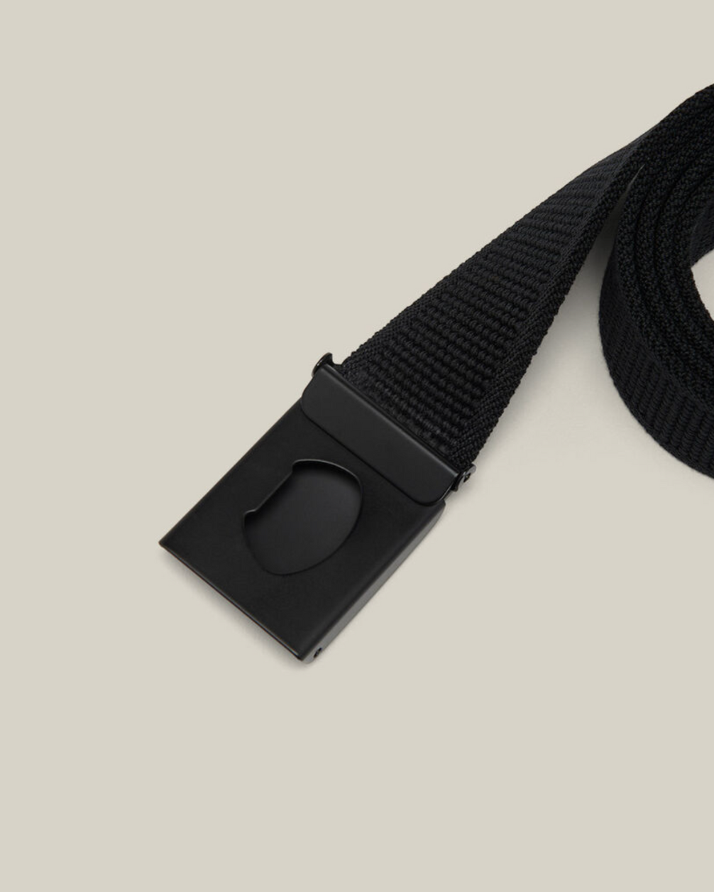 A close-up image of the Hard Yakka Stretch Webbing Belt, showcasing its black woven fabric and rectangular black metal buckle. The smooth, matte-finish buckle features a distinctive cutout detail. The background is a plain light beige color.