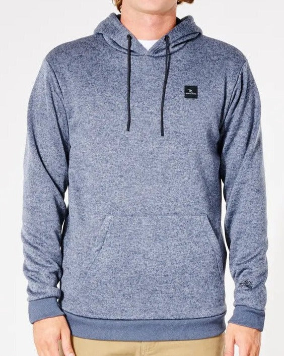 A person stands against a plain background wearing a gray Rip Curl Anti Series Crescent Hood with drawstrings, a small black chest logo, and front pocket. They pair the durable water-resistant hoodie stylishly with beige pants.