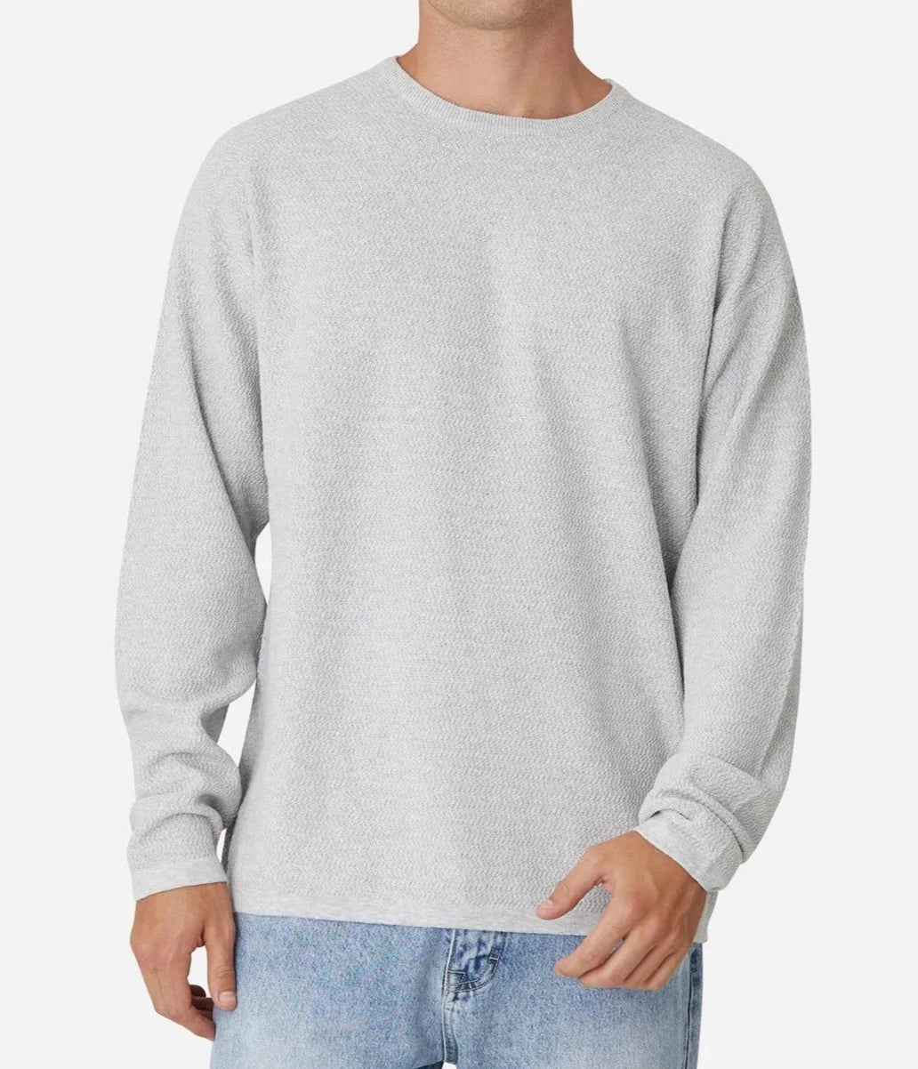 A person wearing the Industrie The Aries Knit in light gray, a 100% cotton long-sleeve crew neck sweater, along with light blue jeans, is standing against a plain white background. The comfortable knit sweater has an oversized fit, giving off a relaxed vibe. The individual is casually resting one hand at their side and the other near the bottom edge of the sweater. Their face is not visible.