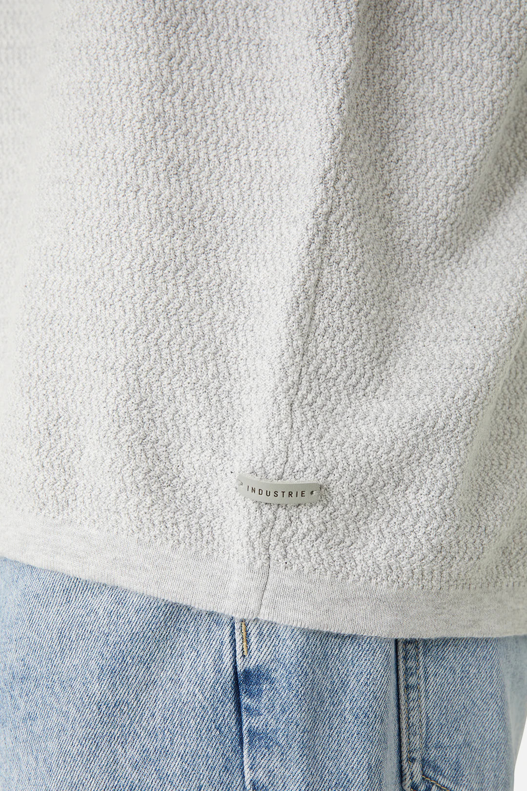 A close-up of a person wearing a textured, light gray garment, likely an oversized fit 100% cotton sweater called "Industrie The Aries Knit," and light blue denim jeans. The metallic tag attached near the hem of the sweater reads "INDUSTRIE." The focus is on the texture and brand detail of the comfortable knit clothing.