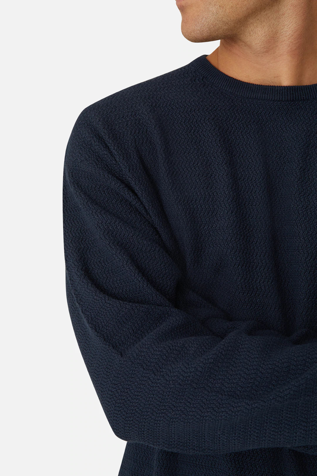 A person in an oversized navy blue textured sweater, named "Industrie The Aries Knit" and made from 100% cotton, stands with arms crossed. They look to the side against a plain light gray backdrop, highlighting their upper body and part of their face.