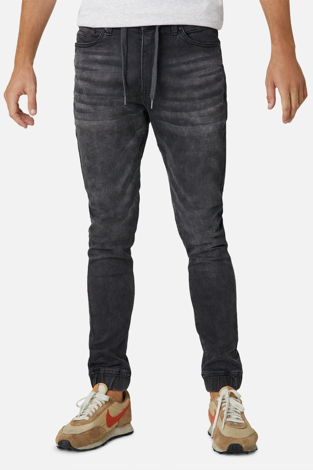 A person wearing a grey T-shirt, Industrie The Drifter Denim Pant in dark grey, which features a jogger style with a drawstring waist, and tan sneakers with a red Nike swoosh stands against a white background. The stylish jeans have a relaxed fit with textured, ribbed designs on the thighs and elastic cuffs at the ankles.