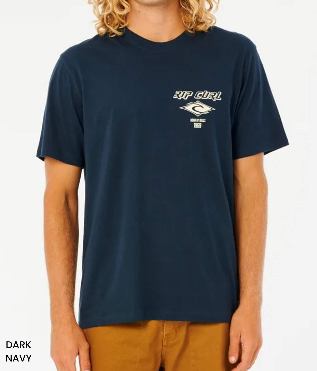 A person wears the dark navy Rip Curl Fade Out Icon Tee, featuring a small left chest logo. Made from organic cotton for eco-friendly comfort, this relaxed fit, short-sleeved shirt with a round neckline pairs well with brown pants against a white backdrop.