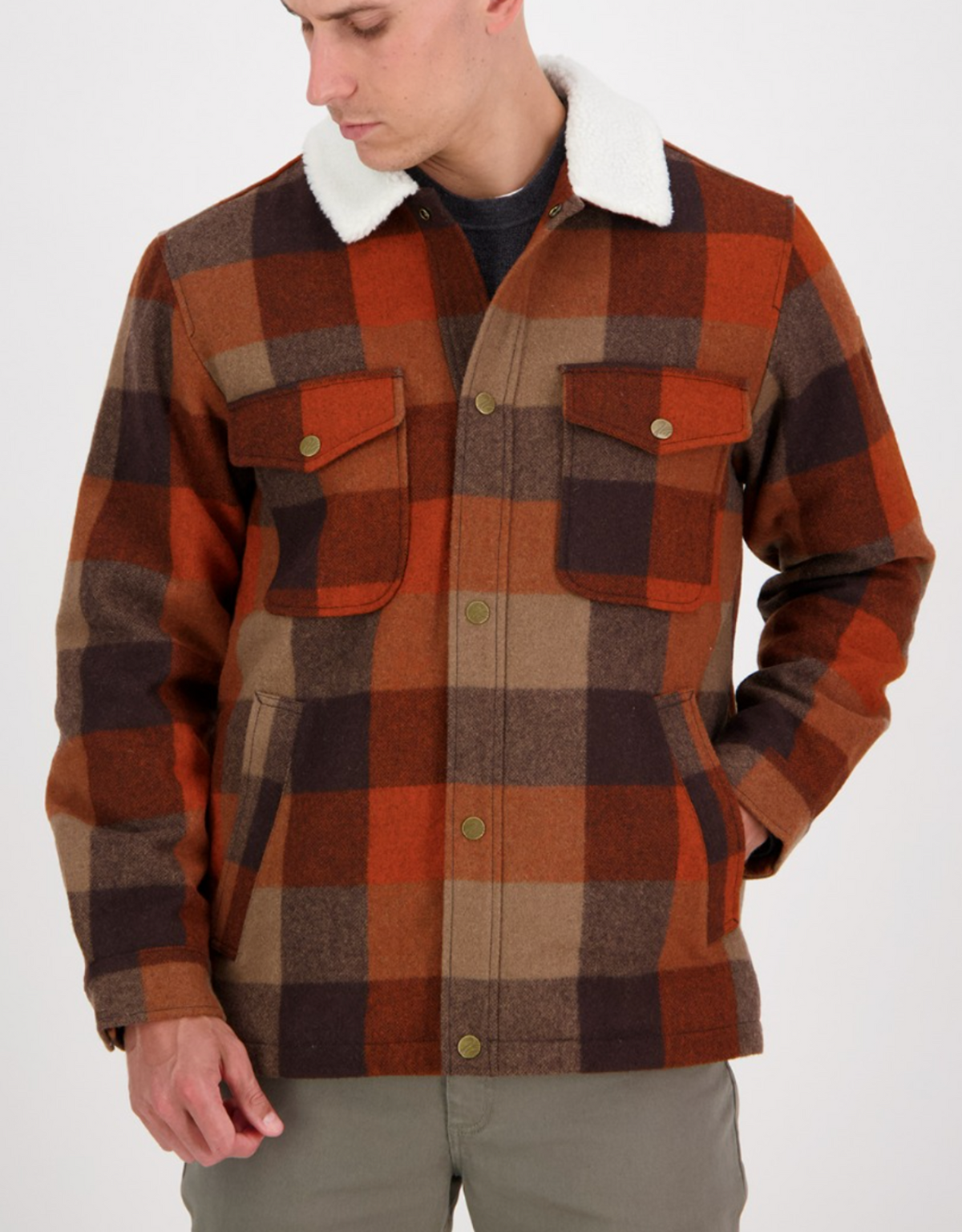 A man wears the Swanndri Kaituna Sherpa Lined Jacket with a red, brown, and beige plaid pattern, white faux-shearling collar, front flap pockets, and snap buttons, looking down to the side with one hand in his pocket.