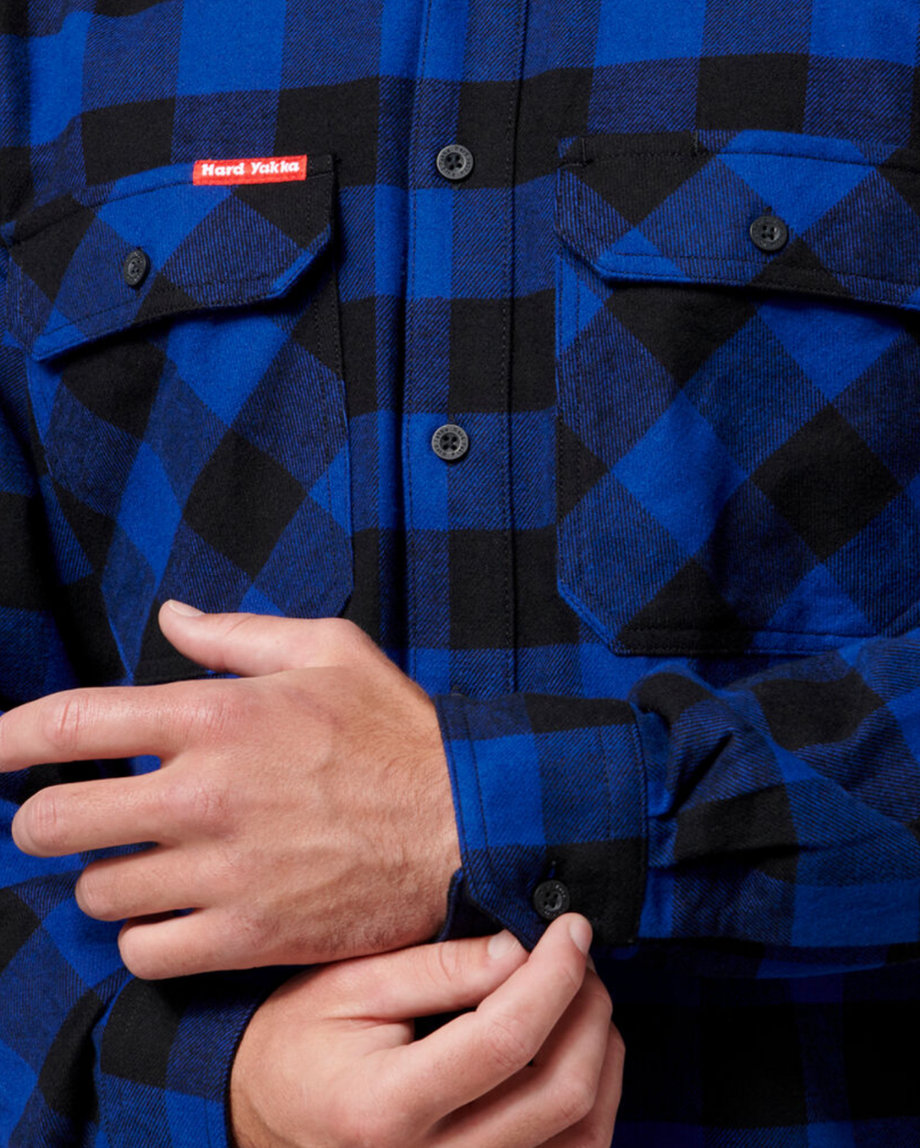 A person is buttoning the cuff of a blue and black plaid, hard-wearing Hard Yakka Check Flannelette Shirt. The Cotton Twill shirt features two front pockets with button flaps and a small red "Hard Yakka" label above the left pocket. The hands are visible, focusing on adjusting the sleeve of this UPF 50+ sun protection garment.