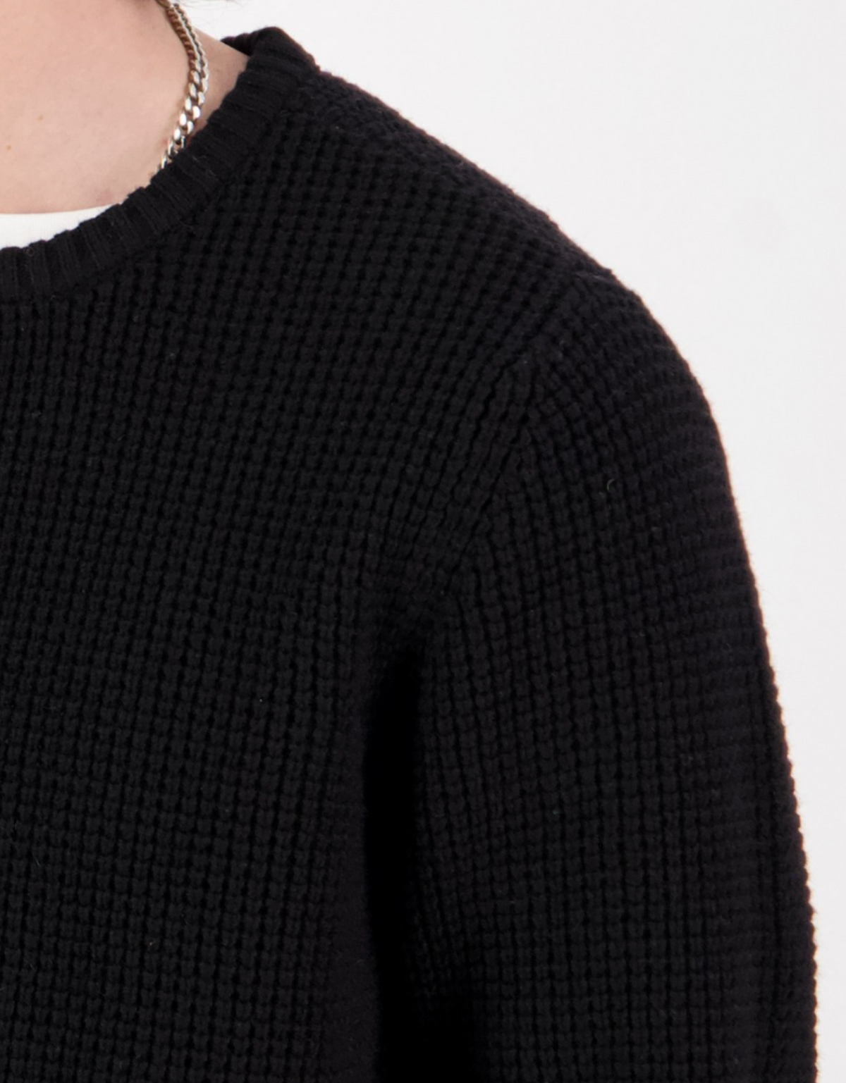 Close-up of a person wearing the Swanndri Fistral Waffle Knit sweater in black, paired with a silver chain necklace and a white shirt peeking out at the neckline, against a plain light background.