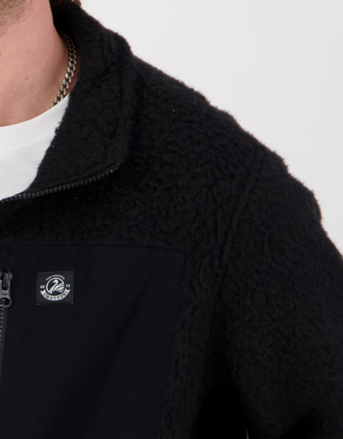 Close-up of a person wearing the Swanndri Churchill Wool Fleece Jacket in black, featuring a logo patch on the chest. The half-zipped design reveals a white shirt underneath, with a chain necklace partially visible around the neck.