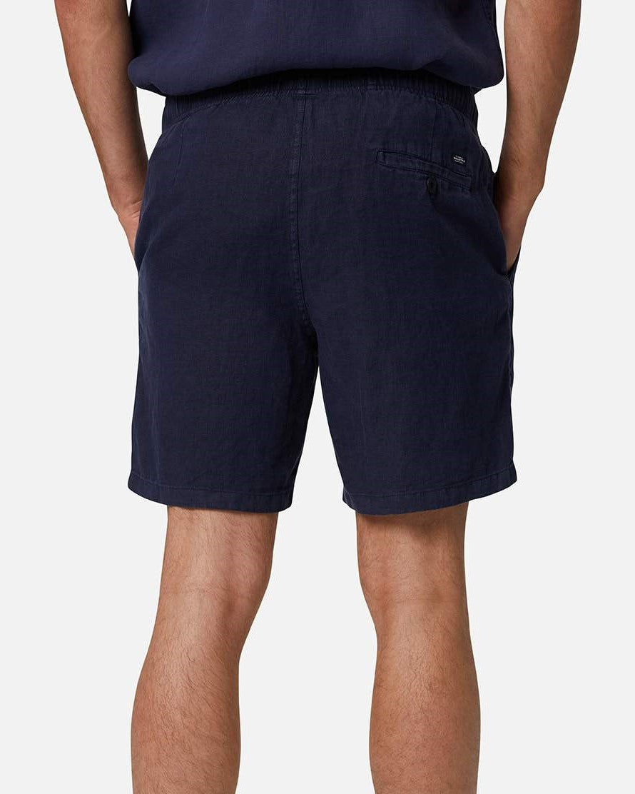 A person wears the Industrie The Marina Linen Short in navy, featuring an elastic drawstring waist and a button-closure back pocket, standing with hands in pockets against a light background.