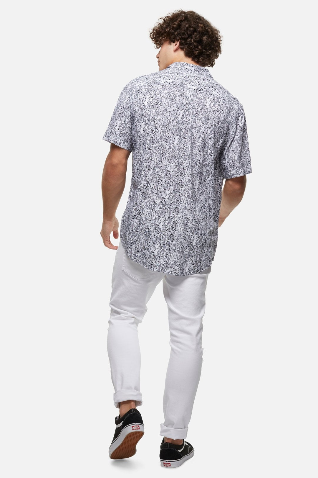 A person with curly hair is seen from the back, wearing a stylish Industrie The Santa Cruz Shirt made of 100% Viscose, white jeans, and black sneakers with white soles and red details. The background is plain white.
