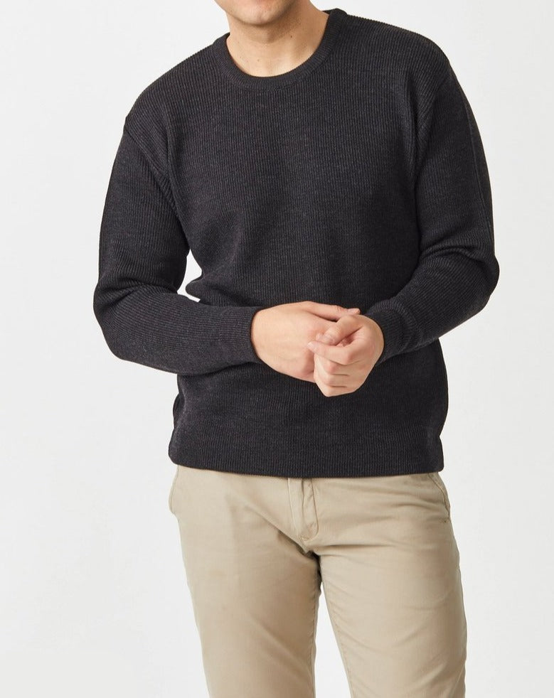 A person wearing a dark Aklanda Fishermans Rib Crew Pullover and beige pants stands against a light background, gently holding their hands in front. The image focuses on the upper body, without showing the face.