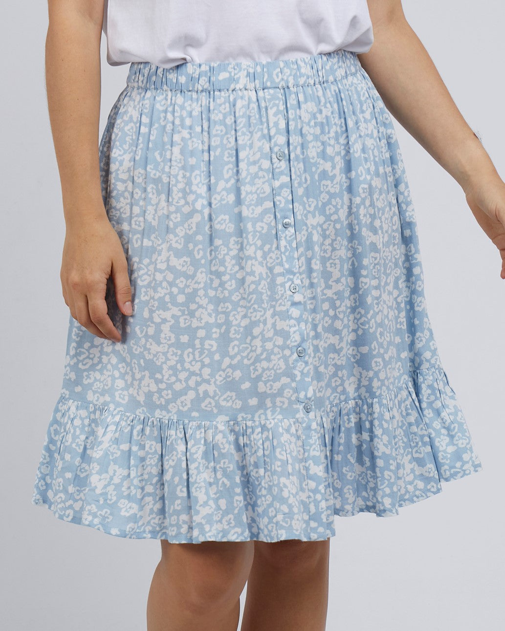 Someone is wearing the Foxwood Kina Skirt, a blue skirt with a white floral pattern and ruffle hem. Made from 100% Viscose, it features front buttons and an elastic waist for comfort. They're also in a white top with their left arm resting by their side.