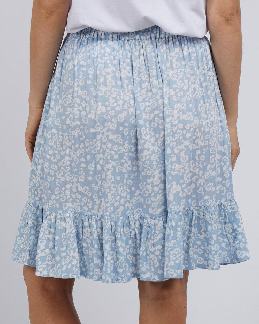A person showcases the back view of a stylish ensemble pairing a white top with the Foxwood Kina Skirt, featuring an elastic waist for comfort, in blue with a light floral pattern and a ruffled hem.