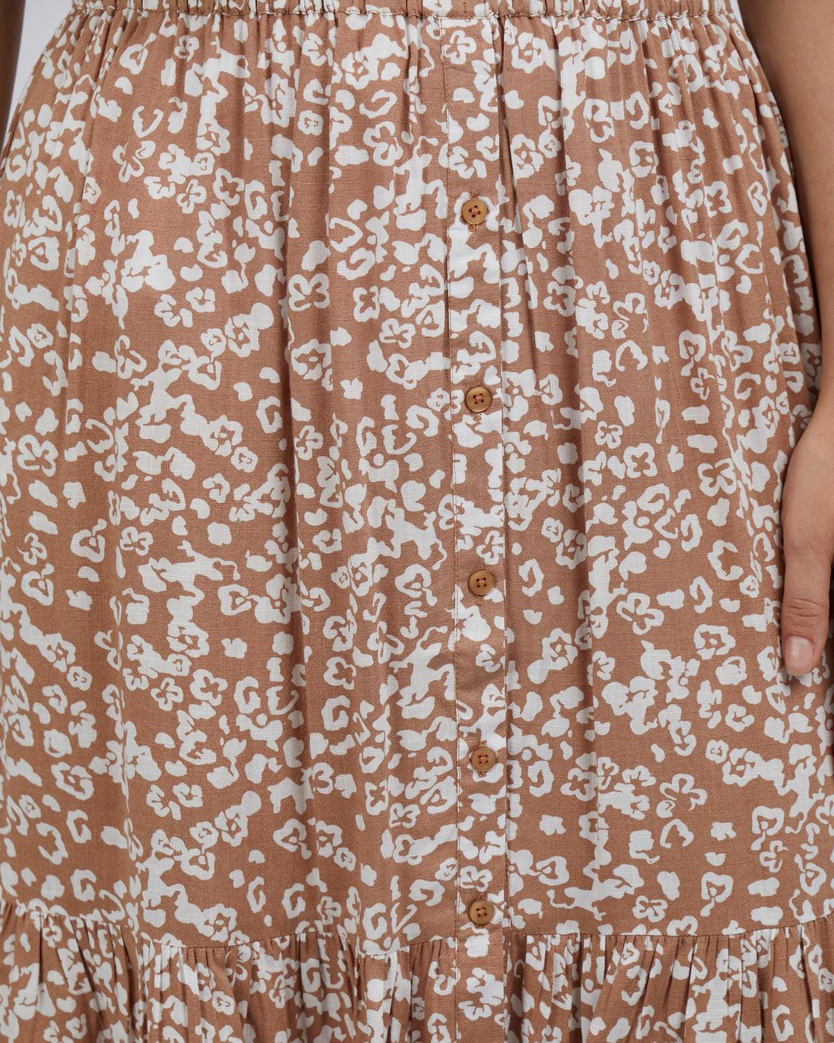 A close-up shows the Foxwood Kina Skirt, a brown piece with a white abstract floral pattern. It features a row of small buttons down the center, gathered details at the elastic waist, and a ruffle hem. A hand gently touches this elegant 100% Viscose garment.