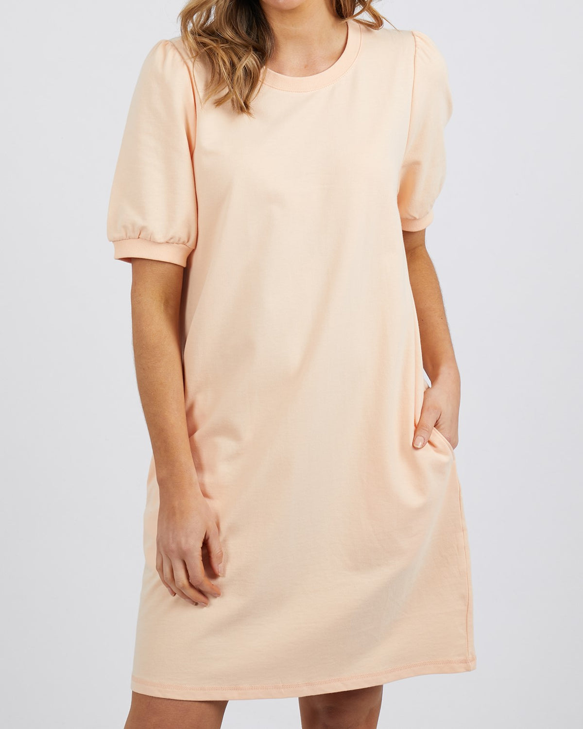 A woman wears the Foxwood Zaliah Dress in light peach, crafted from 100% cotton with handy pockets. Her brown hair falls in loose waves against a plain background. The dress's short sleeves subtly suggest charming puff sleeves, blending style and comfort seamlessly.