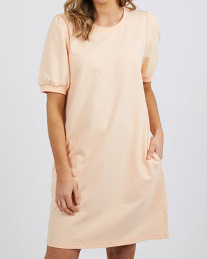 A woman wears the Foxwood Zaliah Dress in light peach, crafted from 100% cotton with handy pockets. Her brown hair falls in loose waves against a plain background. The dress's short sleeves subtly suggest charming puff sleeves, blending style and comfort seamlessly.