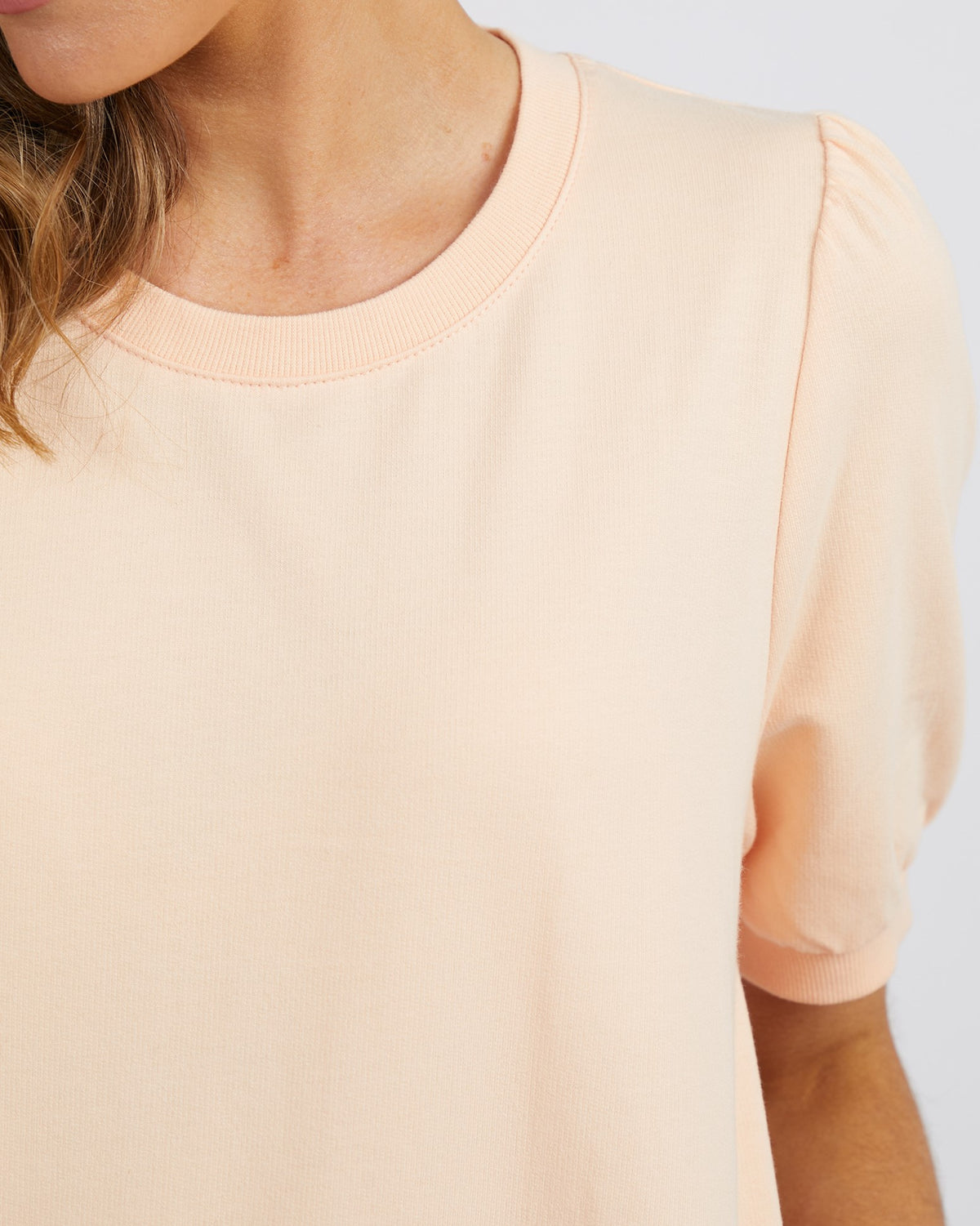 A close-up of a person wearing the Foxwood Zaliah Dress in light peach 100% cotton, featuring short sleeves and a round neckline, with their wavy hair visible on the left side against a plain white background.