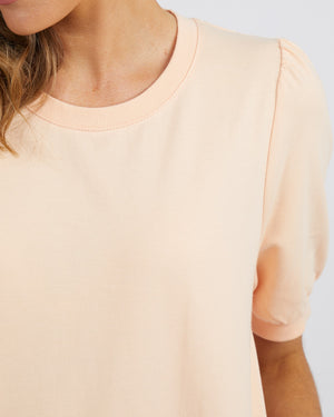 A close-up of a person wearing the Foxwood Zaliah Dress in light peach 100% cotton, featuring short sleeves and a round neckline, with their wavy hair visible on the left side against a plain white background.