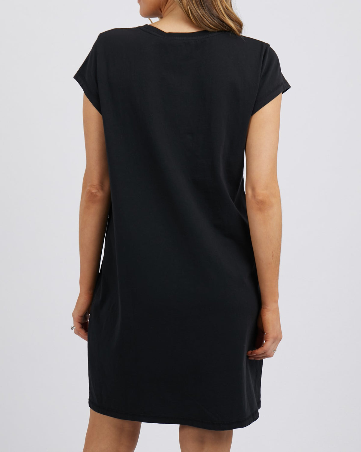 A woman is shown from the back wearing the Foxwood Signature Dress, a simple, short-sleeved, knee-length casual style. Her arms are relaxed at her sides as she stands against a plain white background.