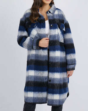 A person wearing a stylish Foxwood Nellie Jacket in blue, white, and black plaid. The long coat features buttons and two chest pockets. They pair it with a white top, black pants, and have long, wavy hair.