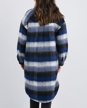 Back view of a person wearing a trendy Foxwood Nellie Jacket, featuring long sleeves and knee-length blue, black, and white plaid stripes. They have long wavy hair and stand against a plain backdrop.