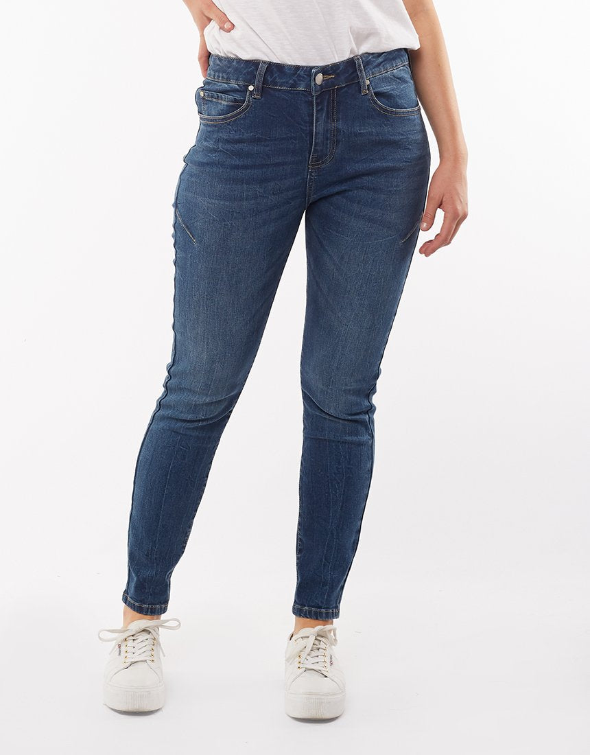 A person stands against a plain white background, wearing a white top, blue denim Foxwood City Jeans, and white sneakers. The high-waist jeans fit snugly with a skinny leg fit and have pockets. The person’s arms are relaxed by their sides and their hands are partially in their pockets.