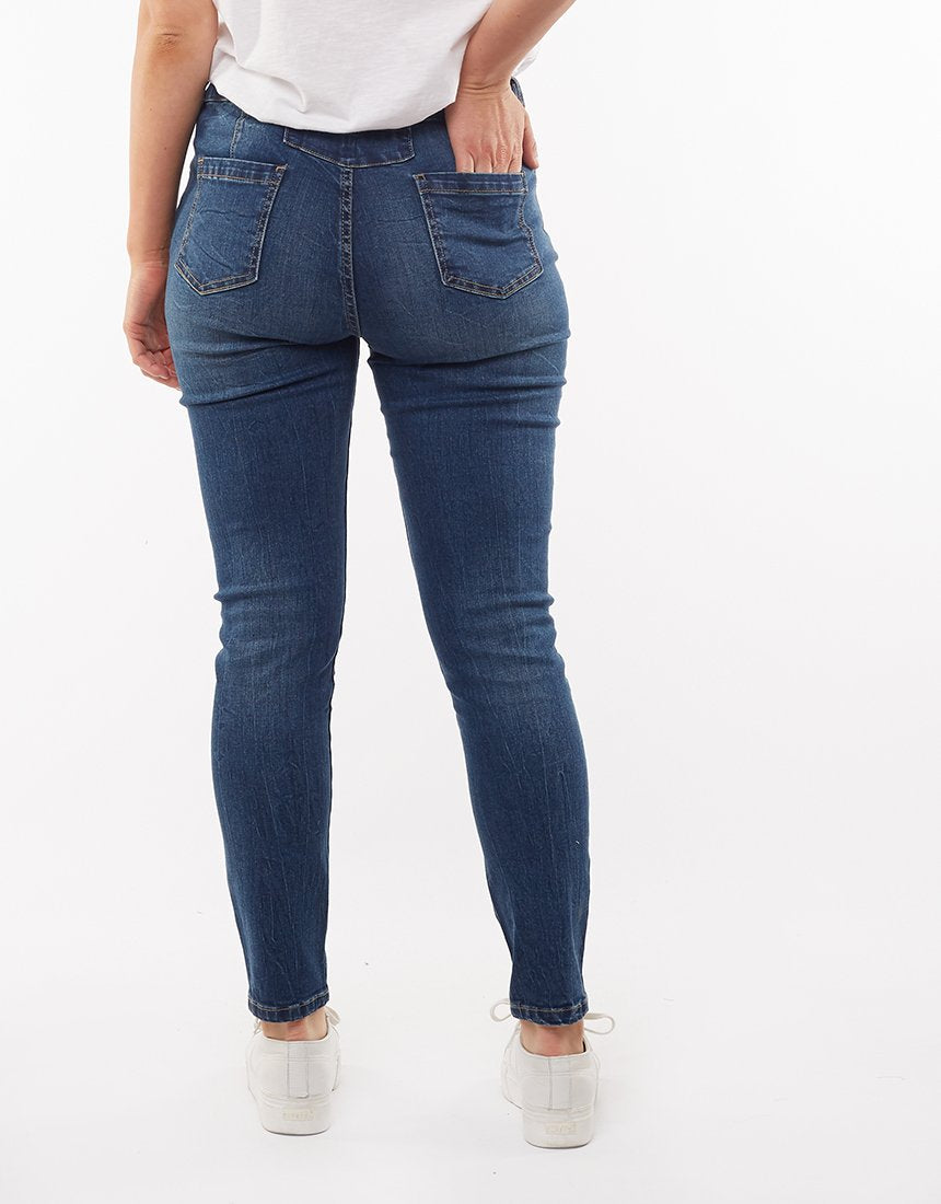 A person is standing with their back to the camera, wearing a white T-shirt, Foxwood City Jean with a high waist and skinny leg fit, and white shoes. One hand is resting in their back pocket. The background is plain white.