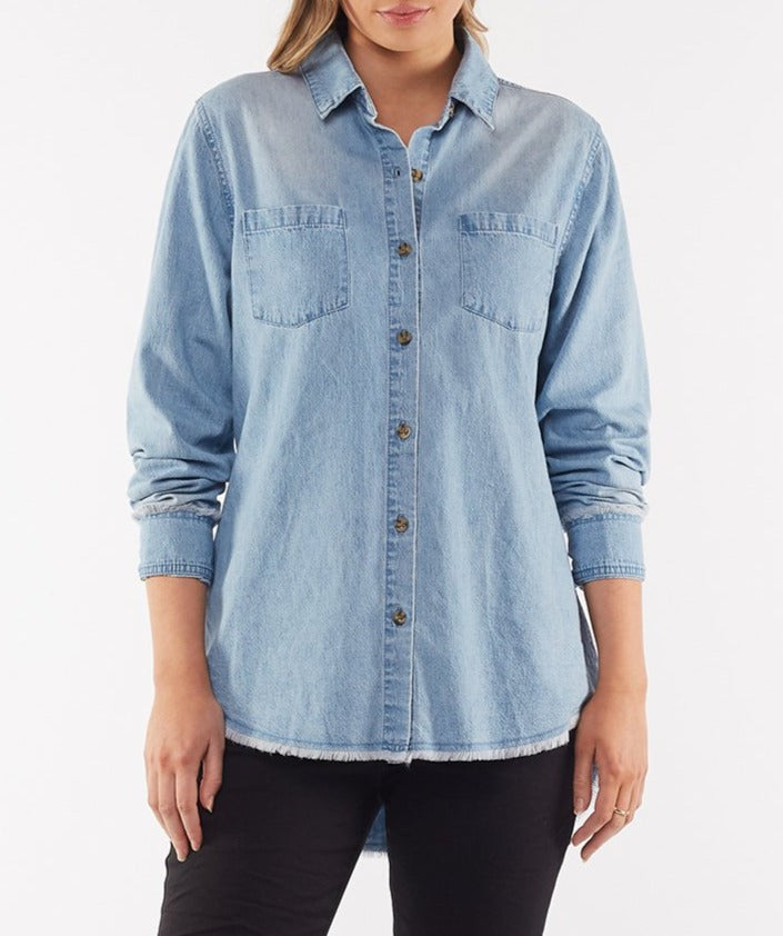 A person wearing the Foxwood Noah Shirt, a light blue denim long sleeve shirt made of sturdy cotton, featuring two chest pockets and a button front. Paired with black pants, only the torso and upper part of the legs are visible against a white background.