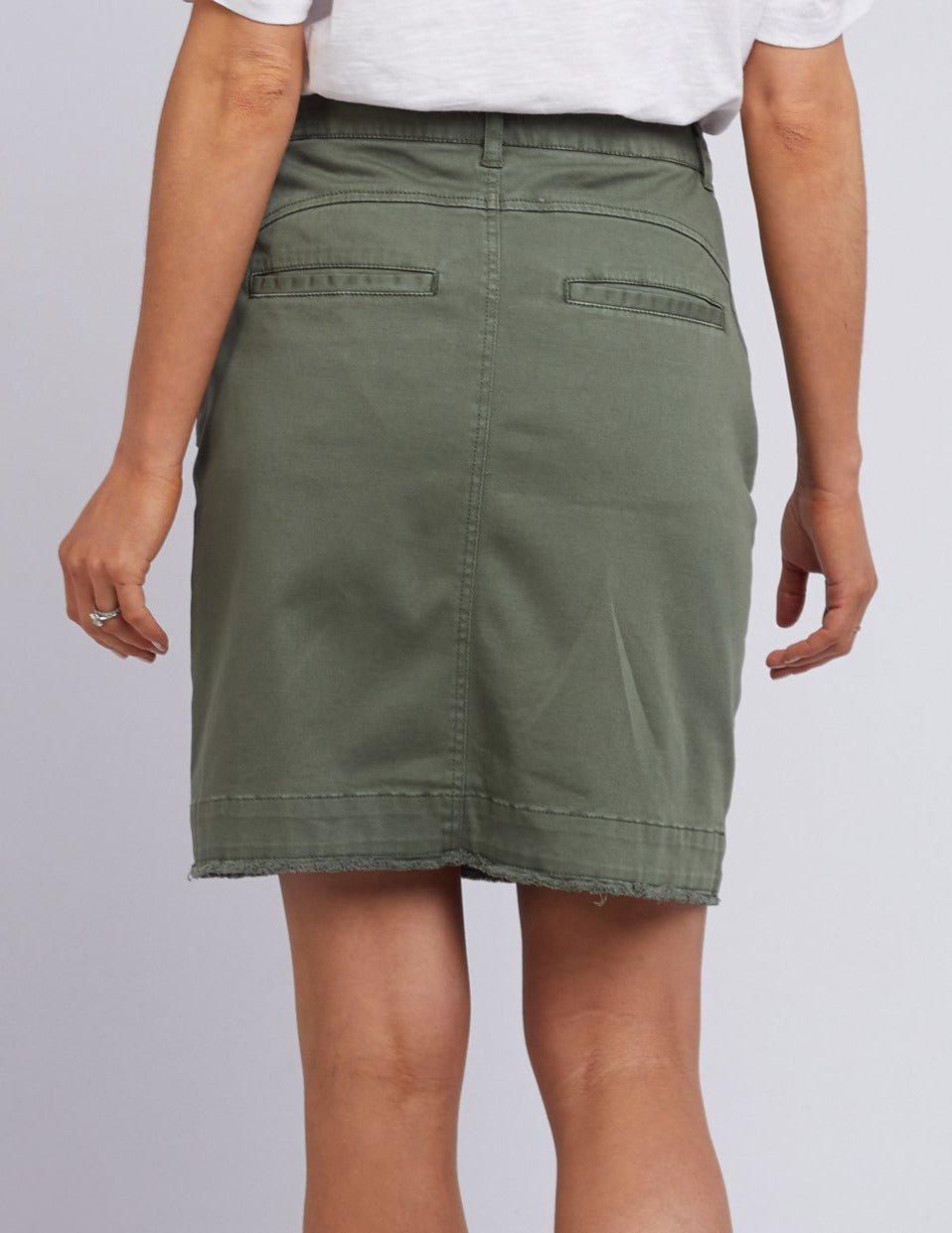 Back view of a person wearing the Foxwood Charlee Skirt in green, made of cotton with a knee-length cut, two back pockets, and a raw hem. They also have on a white top. The right hand is shown with a small ring on their finger.