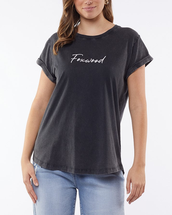 A person confidently wears rolled-up short sleeves of a "Foxwood Signature Tee" in black, crafted from lightweight, breathable fabric. They pair it with light blue jeans and stand against a plain white backdrop, one hand on their hip.