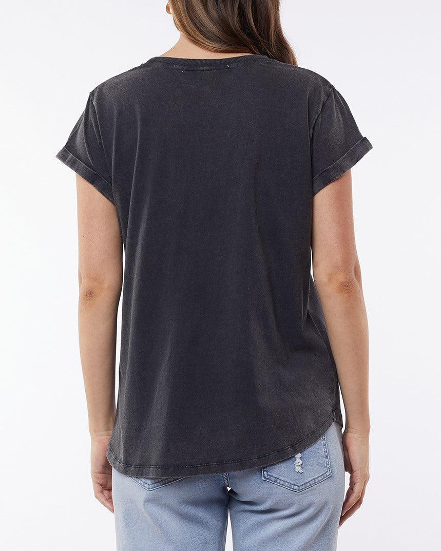 View of a person with long hair wearing the Foxwood Signature Tee, a faded black T-shirt made from lightweight breathable material, paired with light blue jeans. The background is plain.