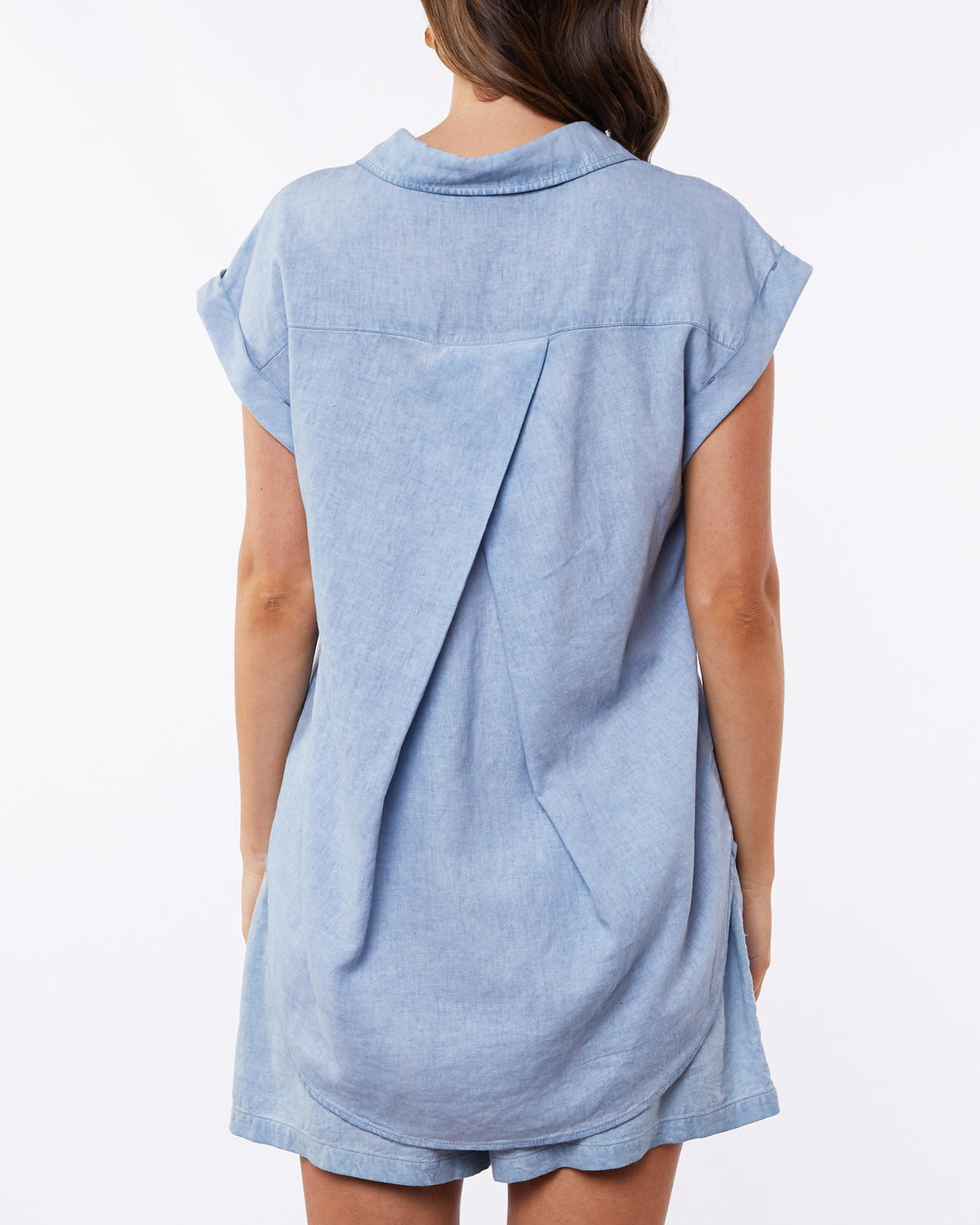 The image showcases the Foxwood Palm Shirt, a light blue short-sleeve denim dress with a relaxed fit and pleated back detail, falling just below the knees. The stylish piece pops against a plain white background.